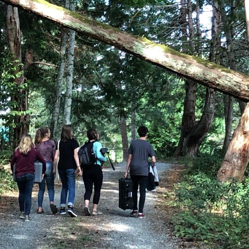 group of five youth walking down dirt road in the forest, carrying film equipment
